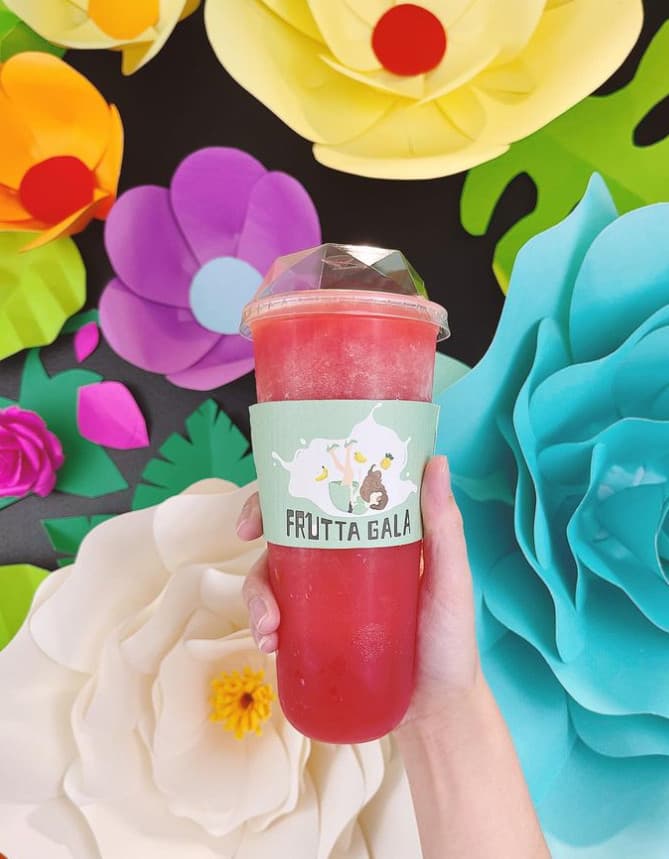 A hand holding a Frutta Gala fruit slush in front of a wall of colourful paper flowers