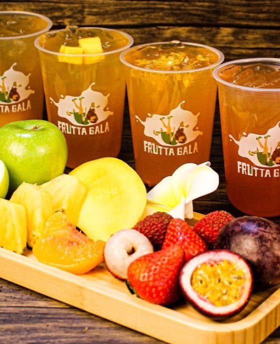 Four iced fruit teas in Frutta Gala cups beside a wooden tray of fresh pineapple, apple, lychee and passion fruit